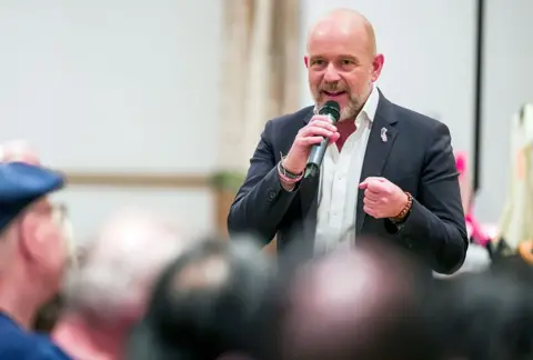 Getty Images Steve Hilton wears a dark blazer and a white button down shirt with no tie. He holds a microphone and speaks at a campaign event.