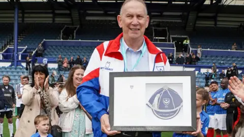 A man with very short hair and wearing glasses on top of his head holding up a framed honour. He's wearing a blue, red and white jacket and smiling at the camera. Behind him, crowds of people are clapping in front of blue raked seating.