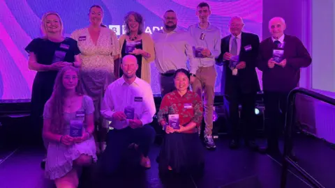 The winners photographed in a group at the ceremony, all smiling and smartly dressed holding their glass awards. Three of them are kneeling in front of a row of winners. They are standing in front of a screen which is purple and has the white BBC Radio Cornwall logo on it.
