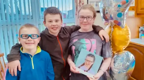 Jennifer Doey Lyla is pictured with her two brothers. She is wearing a black and pink T-shirt and is wearing circled glasses. Her brother Simeon 