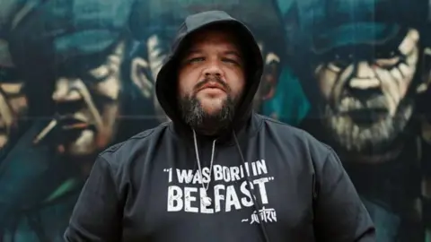 Skeet/Peter Whelan Jun Tzu. He has a dark beard, wearing a black hoodie with the hood up, on the front of it it reads 'I Was Born In Belfast'. A mural of men wearing flat caps and smoking cigarettes is behind him.