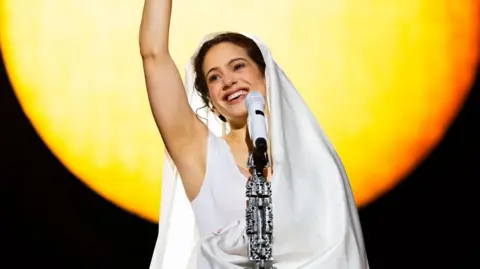 Rosalia on stage in a white shawl in front of a microphone stand, smiling with one arm in the air, in front of a yellow sun video backdrop