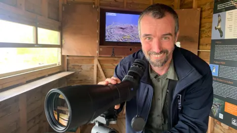 A man wearing a blue coat with the Leicestershire and Rutland Wildlife Trust logo on it. He is holding on to a spotting scope in a bird hide at Rutland Water. The Trust's live web camera feed of the nest can be seen on the television behind him.
