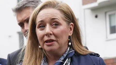 Reuters BBC Spotlight NI reporter Jennifer O'Leary outside the High Court in Dublin. She has long, straight auburn hair and is wearing a navy trench coat over a white blouse. She is also wearing a multi-coloured blue and white scarf.