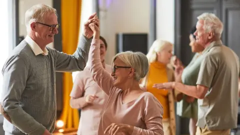 Getty Images Two older adults dancing together in a bright indoor space, with one raising the other’s hand, while other older adults socialise and dance in the background.