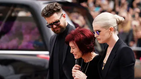 PA Media From left, Jack, Sharon and Kelly Osbourne stand alongside Ozzy's funeral hearse, as they look at the floral tributes laid in the day's after the Black Sabbath frontman died, in July.