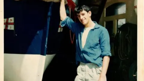 Richard Spindler Richard when he was younger wearing a blue shirt and beige trousers leaning against an old RNLI boat smiling at the camera.