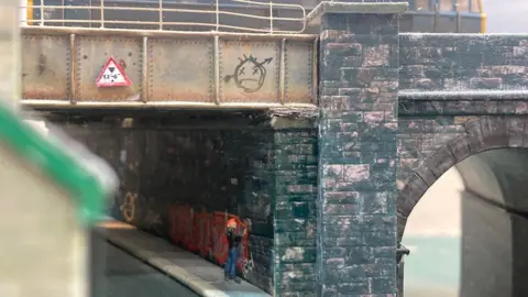 A miniature model of a man with a red Mohican haircut in a black leather jacket and blue jeans spray painting a railway bridge way