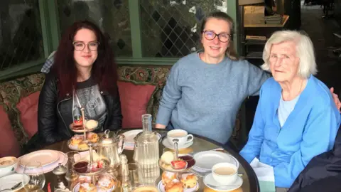 (l-r) Granddaughter Ciara, mother Janie Davies and Grandmother Nora all smiling at the camera. The women are sitting at a table with afternoon tea in front of them to celebrate Nora's 93rd birthday