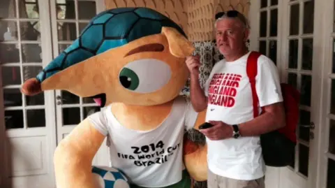 Dave Thompson The image shows a man posing with a World Cup Mascot that is an armadillo. It is wearing a top that says "2014 Fifa World Cup Brazil". The man is wearing a top that says England in a red font.