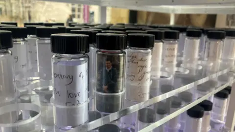 A close-up photograph of the NHS blood contamination scandal memorial. The image shows blood vials, with hand-written personal messages inside. 