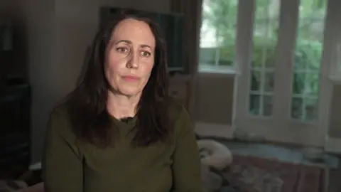 A woman with brown hair and a brown jumper. She is sitting inside what looks like a living room in a family home.