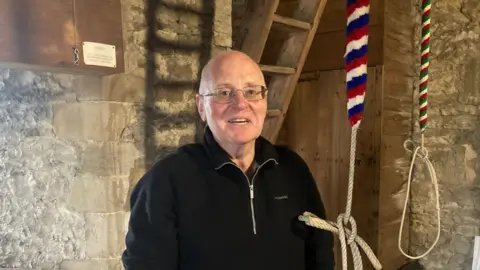 A man stands in a traditional bell‑ringing chamber made of stone and wood. The room has thick stone walls with a rough, aged texture, suggesting an old building such as a church tower or heritage structure.