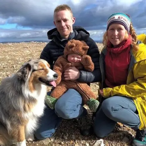 Morag Skelton A family picture with Hamish in a baby suit resembling a brown bear, being held by his father. Morag is on the right and their dog is on the left.