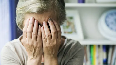 Getty Images An elderly woman with white hair holds her face in her hands