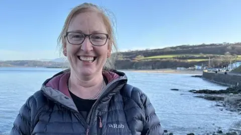 Woman wearing a dark blue down jacket with a purple neck lining, with Benllech beach and sea behind her. She has red hair, and is wearing glasses.