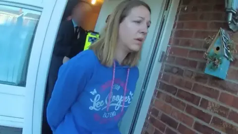 PA Media Lucy Letby, wearing a blue hooded top, is led from the front door of her home.