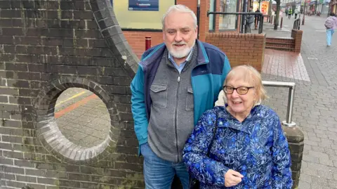 BBC A man and woman stand beside a circular, purpose-made hole in a wall on a town high street. The man has grey hair and a beard, wearing a grey zipped top, green waterproof coat and jeans. The woman has short blonde hair, black glasses and a blue waterproof coat.