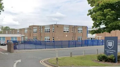 The main entrance to the school with a blue sign saying Co-op Academy Bebington and a two storey school building.