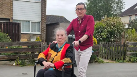 A man in a red shirt and white jeans standing behind an elderly woman in a wheelchair. She is wearing a gold, red and black jacket over a blue top. They are both outside a house while looking into the camera.