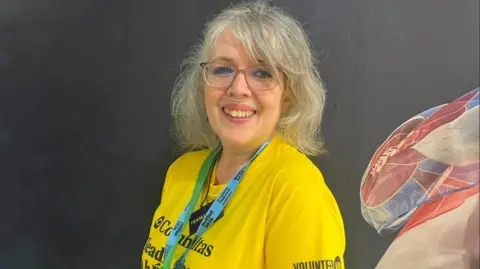 Volunteer now A woman, Camilla Reynolds pictured In front of a black wall with blue, white and red wall art on the right corner. She has grey hair and is wearing a yellow t-shirt with a blue lanyard and is wearing red glasses. 