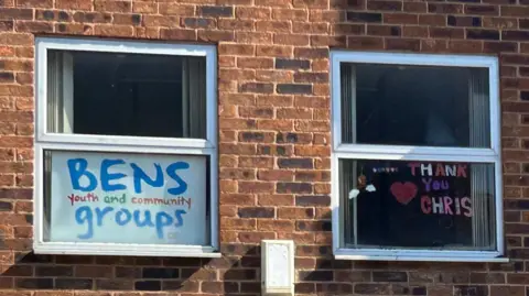 Gavin Kermack A close-up of two windows in a brick wall. One has a sign in reading "BENS Youth and Community Groups". The other has been decorated with a heart and letters reading "THANK YOU CHRIS".