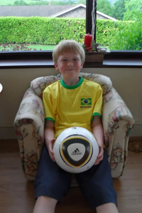 Ieuan Phillips 10-year-old Ieuan sits cantered in a padded armchair, holding a full-size white Adidas football in his lap with both hands. He has short blond hair.  The ball features bold black and gold curved panels and the Adidas logo clearly visible at the front, making it the focal point of the image. The child is wearing a bright yellow Brazil football shirt with green trim and a crest reading “Brasil,” visually linking the clothing to the sport