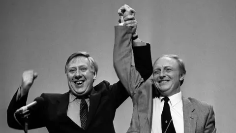 PA Media A black and white image of two men in suits holding each other's arms in the air
