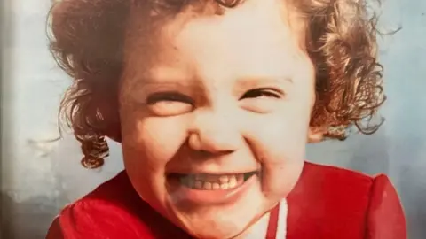 A happy young Katrice smiles at the camera wearing a red dress. She was two when she went missing