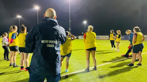 A man wearing a dark coat with Neurodiversity Coach on the back standing behind an all-female team on a well-lit pitch