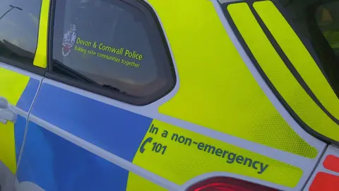 The side of a Devon and Cornwall Police car. The car is bright yellow and blue and has 'Devon and Cornwall Police Building safer communities together' written on the rear window.