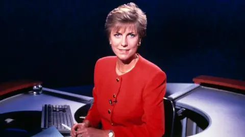 Jill Dando sitting behind a newsdesk on a television set for the 9 o'clock news in 1992. She is wearing a red suit and has short blonde hair.