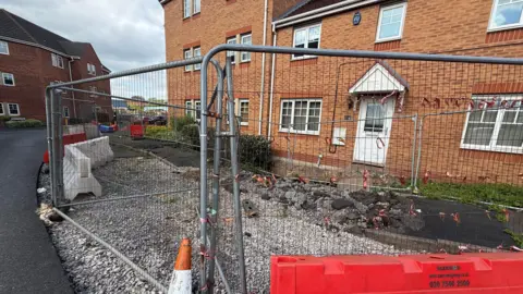 Metal fencing surrounding the front of a row of houses, with a pile of gravel and chunks of broken pavement in front of it. The houses are brick-built, with white window frames and door. There are small pieces of red fabric attached to the fencing.