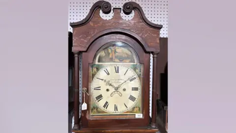 Chilcotts Auctioneers and Valuers A brown wooden clock with a painted face that depicts houses and green grass and a boy above the face. The picture of the clock is on a light purple background. 