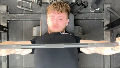 Lucas training his bench press - looking down on him as he performs the movement, holding a barbell above his chest, wearing a black T-shirt and clearly straining as he carries out the lift