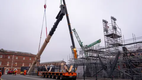 NMRN Scaffolding obscures the dock where HMS Victory sits. On the quayside, its mast is hanging from a large crane.