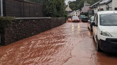 William Westgate A car driving through flood water on a village road 