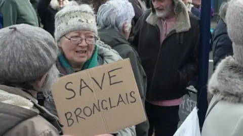 A woman standing amongst a crowd of protestors. She is holding up a cardboard sign with "Save Bronglais" written on. She wears a grey patterned woollen head band, a multi-coloured scarf and a grey jacket.