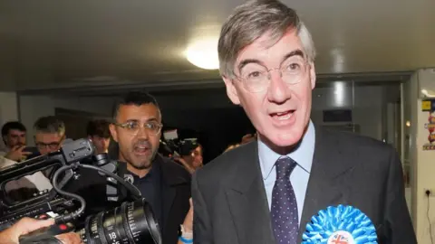 PA Media Jacob Rees-Mogg speaking with a camera next to him and a crowd behind