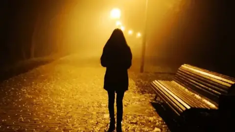 A woman walks along a dark cobbled street with street lights. A bench is to her right.