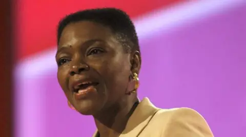 A woman with cropped black hair speaking at a lectern. She is wearing a yellow blazer over an orange t-shirt and gold earrings. She is mid speech, gesticulating to make a point. Behind her is a purple and red backdrop.