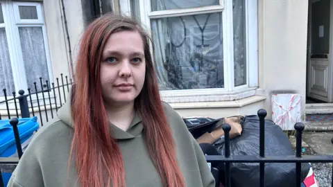 A lady with a glum look on her face stood outside of her old house. She is wearing a green hoodie and has long red hair.