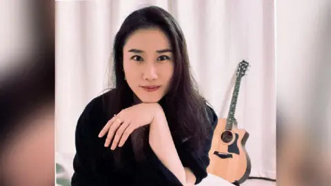 Snow Rui A woman dressed in black sits in front of a white curtain, resting her chin on her wrist. There is an acoustic guitar on a stand in the background.