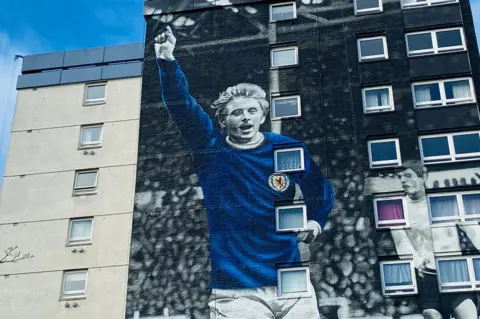 High-rise building with mural of late footballer Denis Law with his arm aloft, in a blue Scotland shirt.