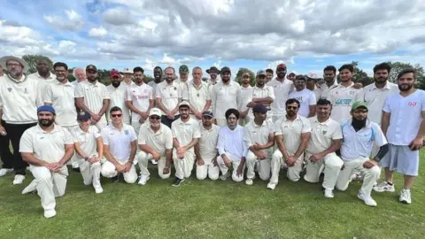 Members of the cricket teams lining up together, wearing cricket whites and smiling at the camera