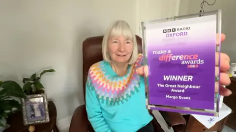 Margo Evans a elderly woman with grey hair sat in the corner of a room in a leather chair. She is wearing a light blue jumper with multi-coloured patterns on it. She is holding a purple glass make a difference award which says that she is the winner of the great neighbour category.