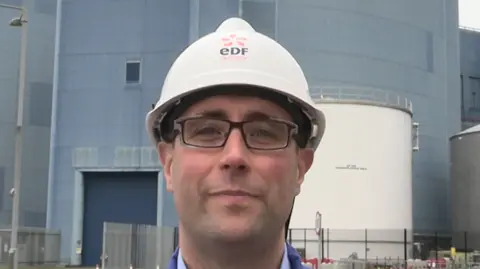 EDF Energy/Christopher Rees Christopher Rees smiles at the camera while wearing a white hard helmet. He is wearing glasses and a blue shirt. Behind him is the Sizewell nuclear station building which sits behind a gated fence.