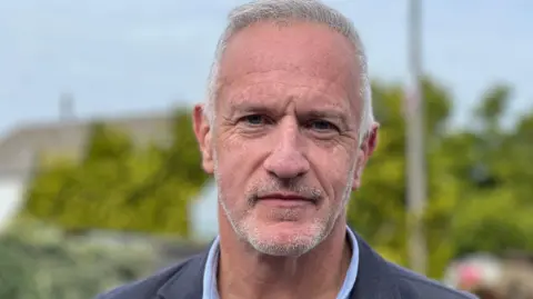 A man with short, grey hair and stubble looks into camera