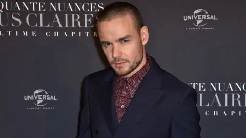 Liam Payne posing for a photograph. He's wearing a dark suit jacket and a maroon shirt. He appears to be at a press event.