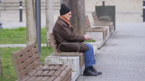PA Media A man is sitting on a bench to the side of a path with grass behind him. He wears a black woolly hat, an orange scarf and brown coat, blue jeans, and black socks and shoes.
